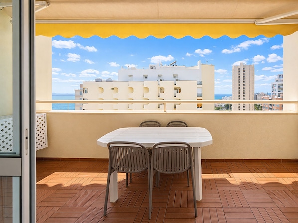 Varanda com mesa branca e duas cadeiras, toldo amarelo e vista para prédios e o mar.