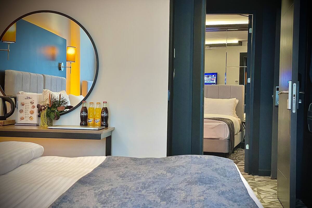 A hotel room with a bed, a desk with a mirror, a small table with a menu and bottles, and a door leading to another room.