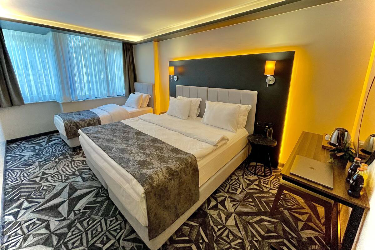 A hotel room with two beds, a patterned floor, a desk with a laptop, and a nightstand with a lamp and bottles.