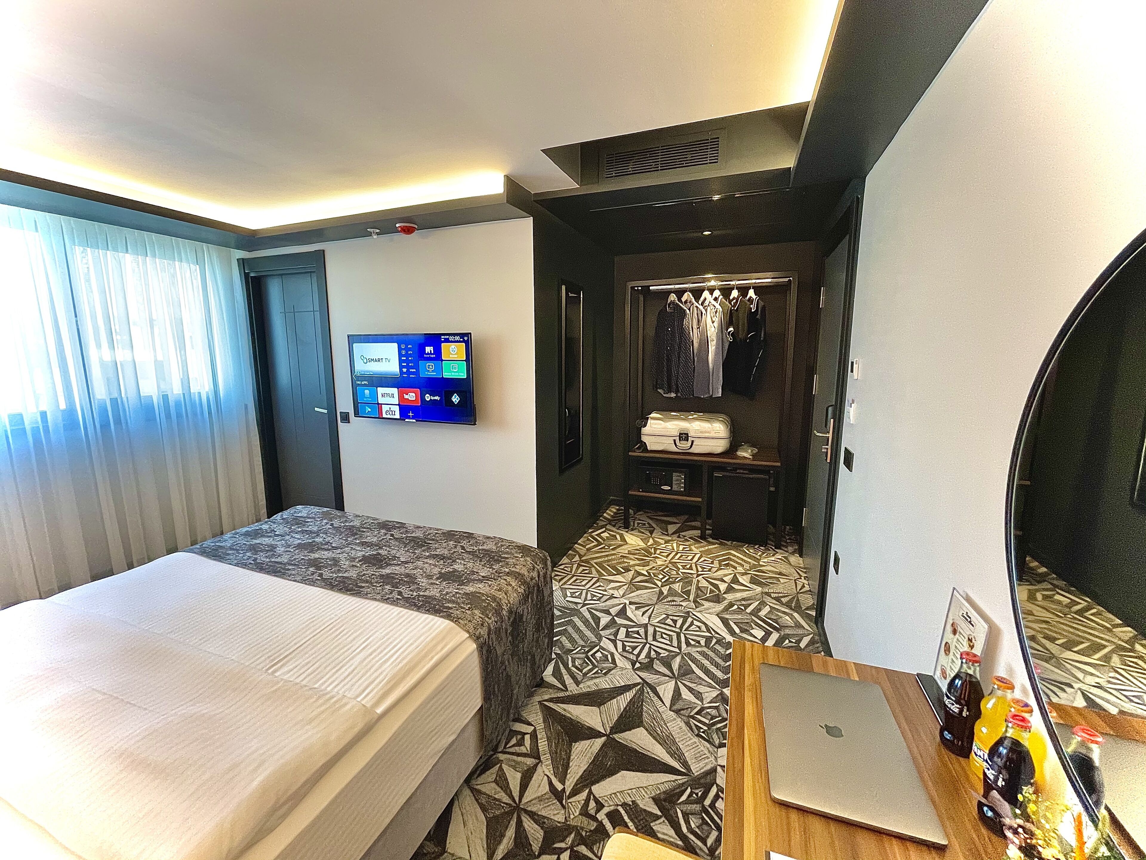 Habitación de hotel con dos camas, un televisor montado en la pared, un escritorio con una computadora portátil y una pequeña zona de estar con un tapete estampado.
