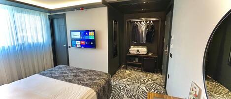 Habitación de hotel con dos camas, un televisor montado en la pared, un escritorio con una computadora portátil y una pequeña zona de estar con un tapete estampado.