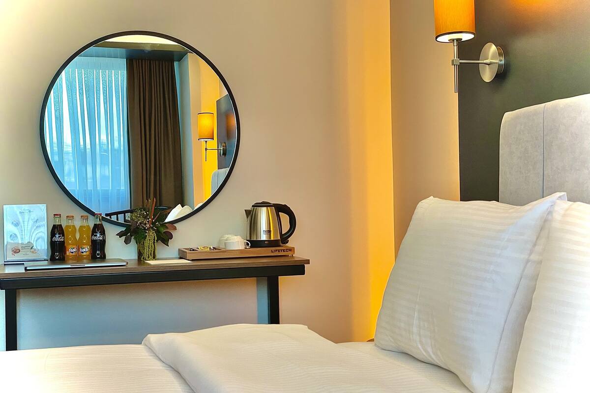 A hotel room with a bed, a round mirror, a desk with a coffee pot, and a wall-mounted lamp.