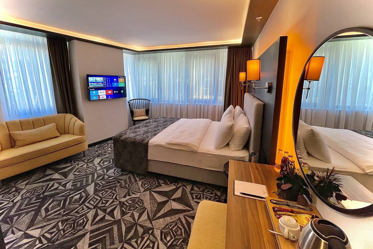 A hotel room with a bed, a sofa, a desk with a mirror, a TV, and a patterned carpet.
