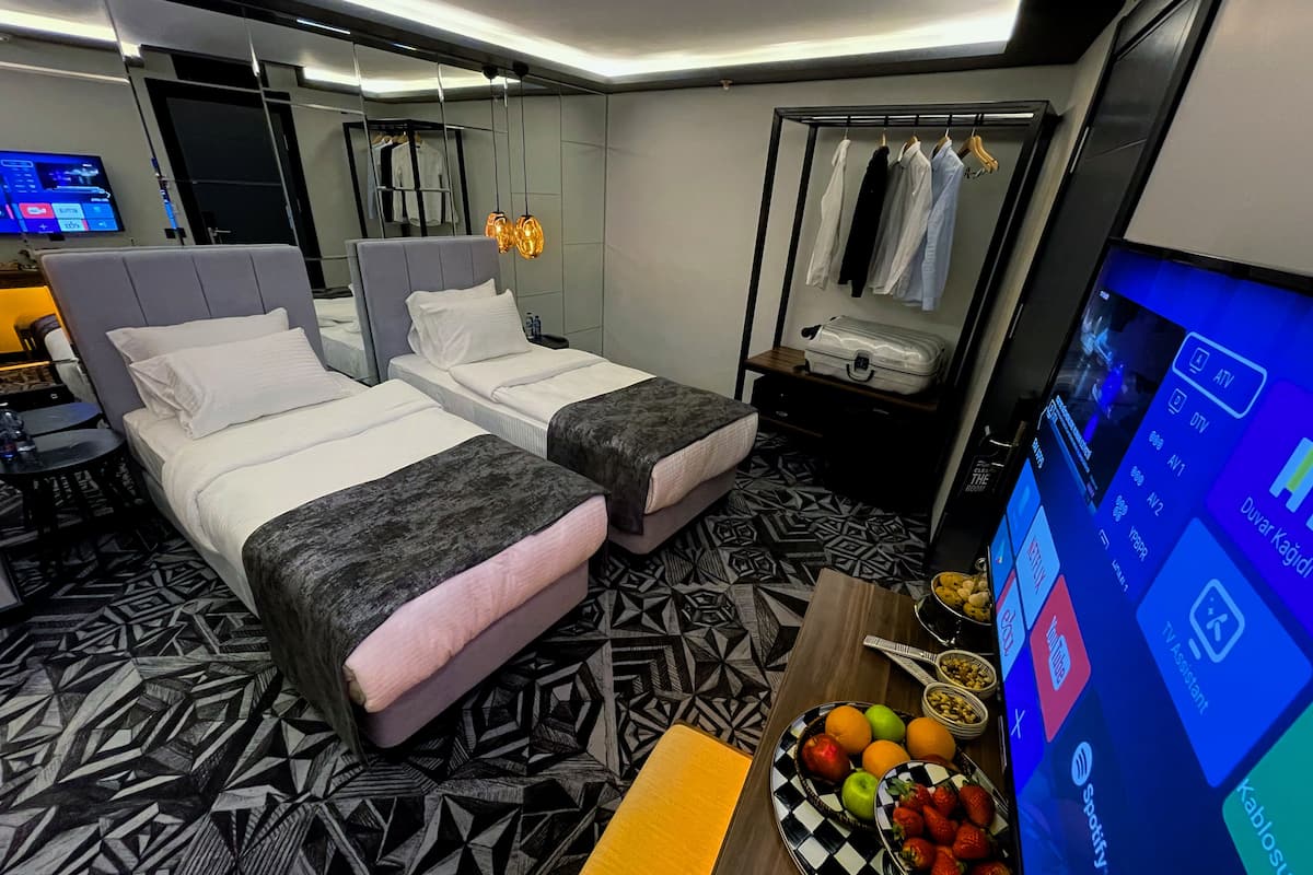 A hotel room with two beds, a TV, a wardrobe, and a desk with a fruit bowl.