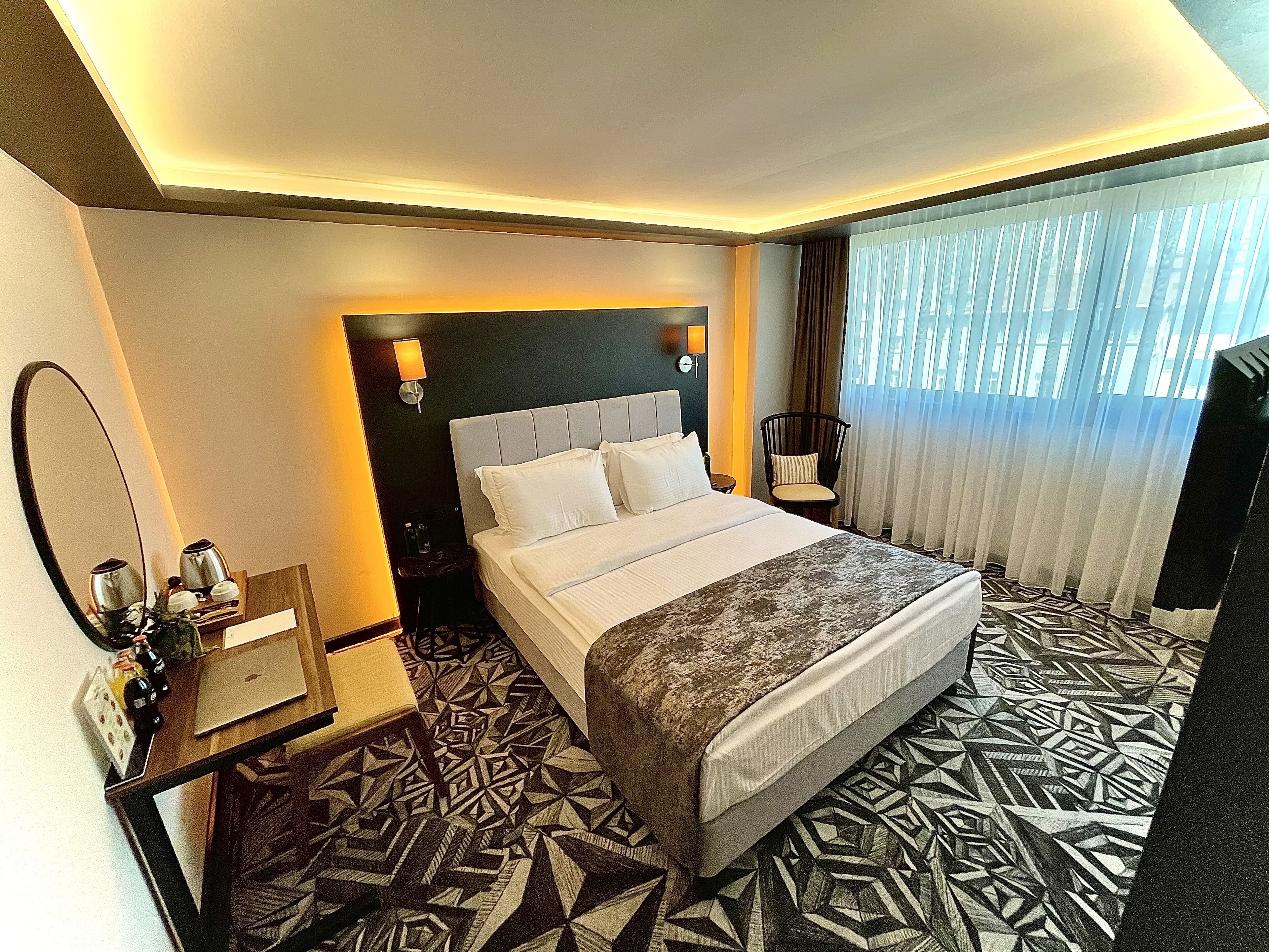 A hotel room with a large bed, a desk with a laptop, a chair, a mirror, and a window with curtains.