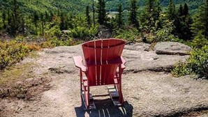 Uma cadeira Adirondack vermelha sobre um afloramento rochoso com vista para um vale florestado e um lago ao longe.