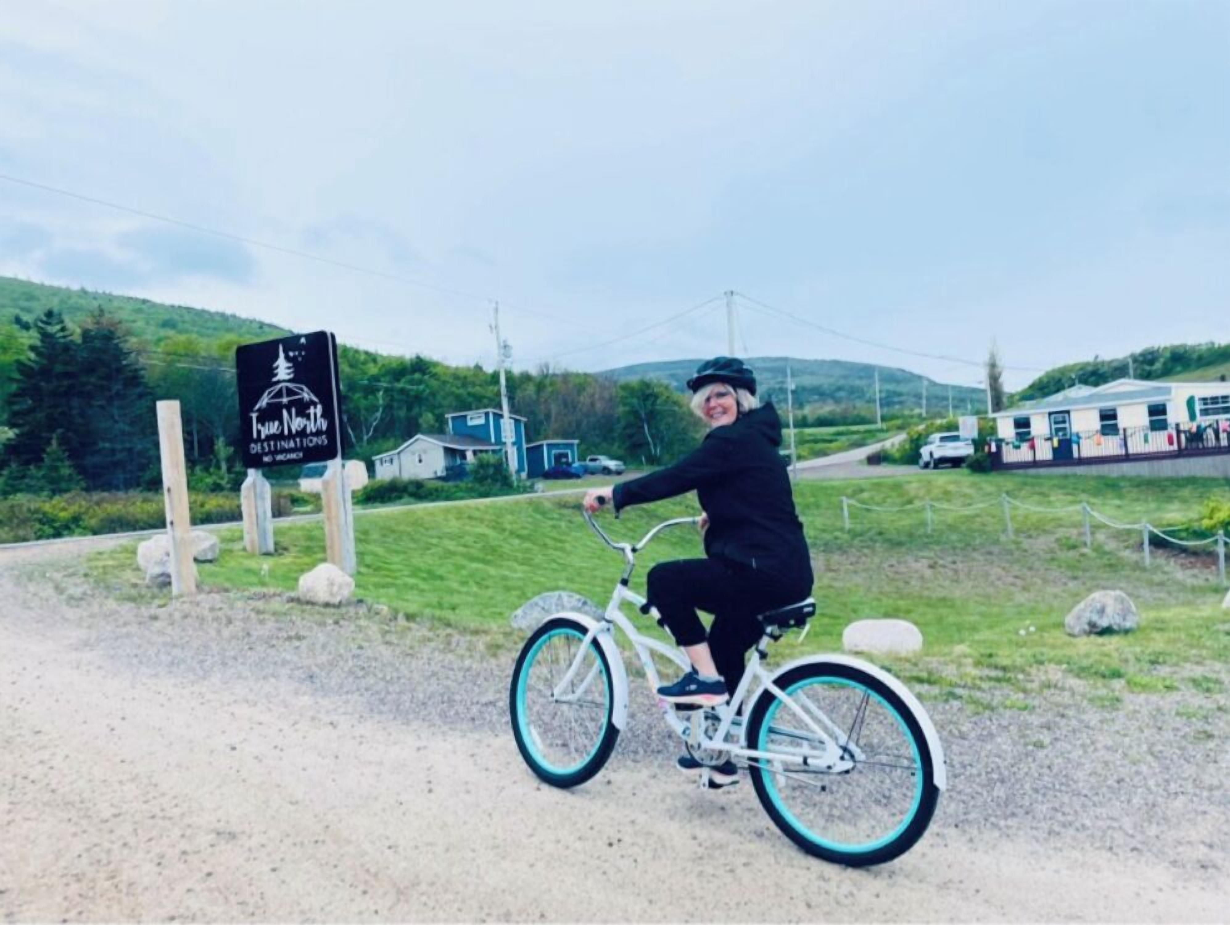 A person riding a bicycle on a gravel path with a sign indicating 'True North Destination' and a small building in the background.