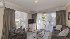 Een ruime woonkamer met een bank, een salontafel en een televisie op een kast. Er zijn grote ramen met gordijnen, een balkon met buitenmeubilair en een plafondlamp.