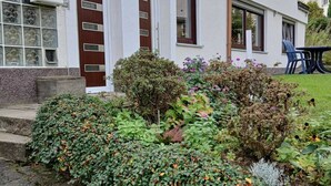 Une maison à deux étages avec une porte d’entrée, un petit porche et un jardin agrémenté de diverses plantes et arbustes.