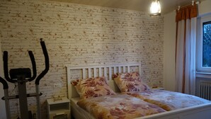 Une chambre avec un mur décoré d’un motif en briques, un lit blanc avec une literie à motifs floraux, une table de chevet et un vélo d’appartement fixé au mur.