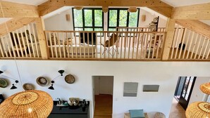 Un loft espacioso con techos altos, vigas de madera, un entrepiso y ventanas amplias que ofrecen vistas a zonas verdes.