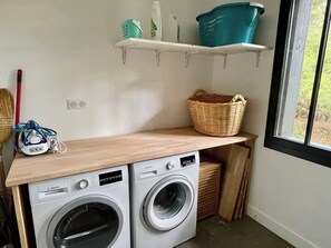 Zona de bugaderia amb dues rentadores de càrrega frontal, un taulell de fusta, un cistell i una prestatgeria amb productes de neteja.