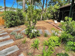 Un jardí amb un pont de fusta, diverses plantes i una construcció petita.