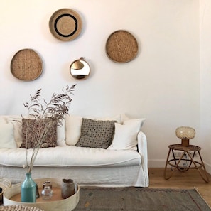 A living room with a white sofa, decorative wall art, a woven stool, and a vase with dried plants.