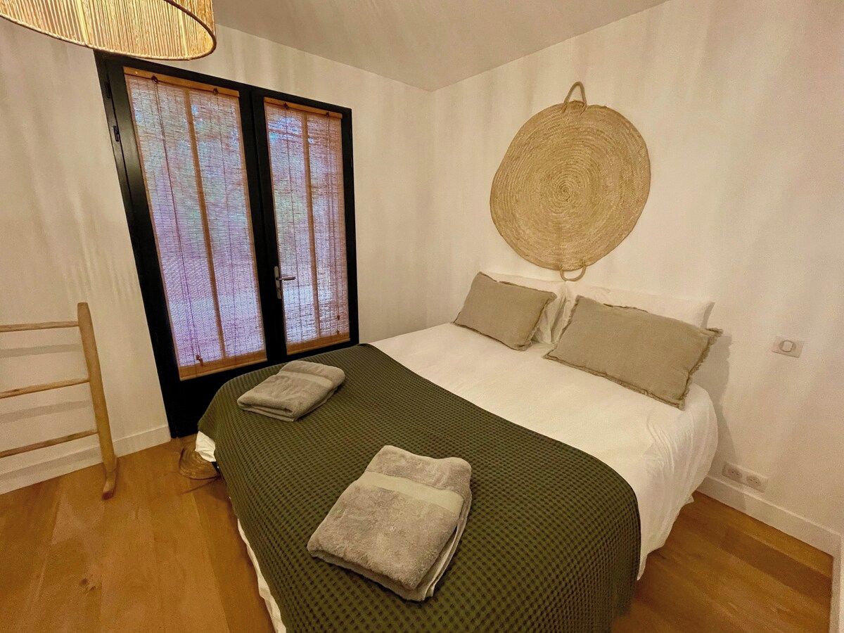 A bedroom with a bed, pillows, a woven wall hanging, a wooden chair, and a window with blinds.