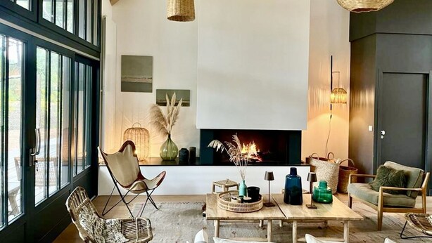 Una sala de estar moderna con chimenea, muebles de mimbre y un ventanal grande.