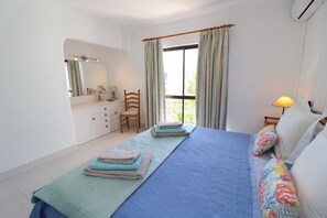 A bedroom with a bed, a chair, a bedside table with a lamp, a window with curtains, and a bathroom with a mirror and sink.