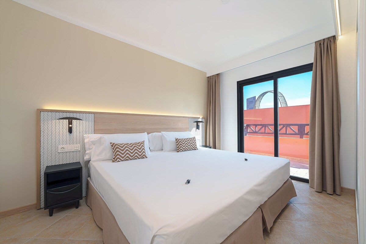 Een hotelkamer met een groot bed, een nachtkastje, een wandlamp en uitzicht op een buitenruimte.