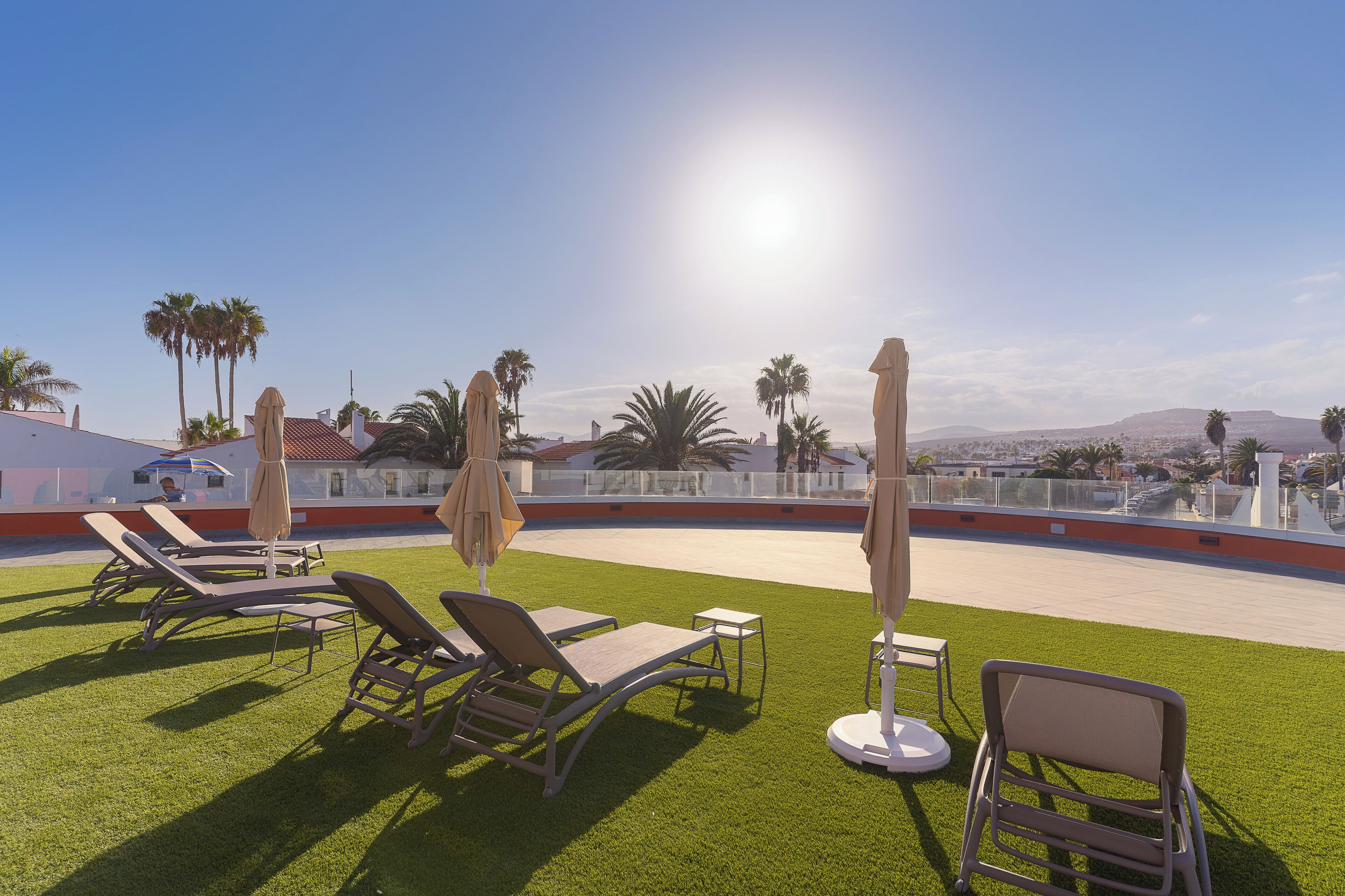 En tagterrasse med liggestole og parasoller, der giver udsigt til palmer og fjerne bakker.
