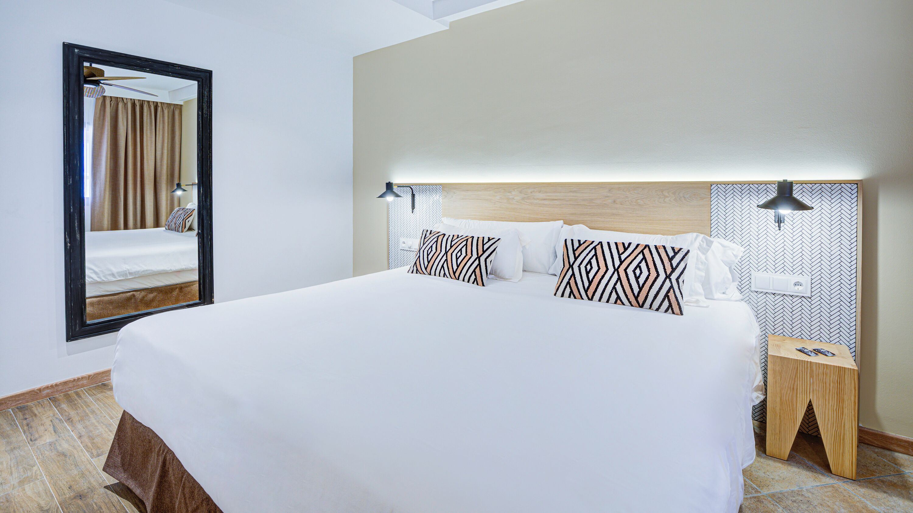 A hotel room with a large bed, a mirror, two bedside tables, and a headboard with built-in lighting.