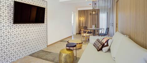 A modern living room with a geometric patterned wall, a flat-screen TV, a sofa with patterned cushions, and a dining area with chairs and a table.