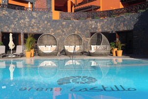 Zona de piscina con tres sillas de mimbre y sillones, un muro de piedra y una piscina cristalina con el texto "arena castillo".