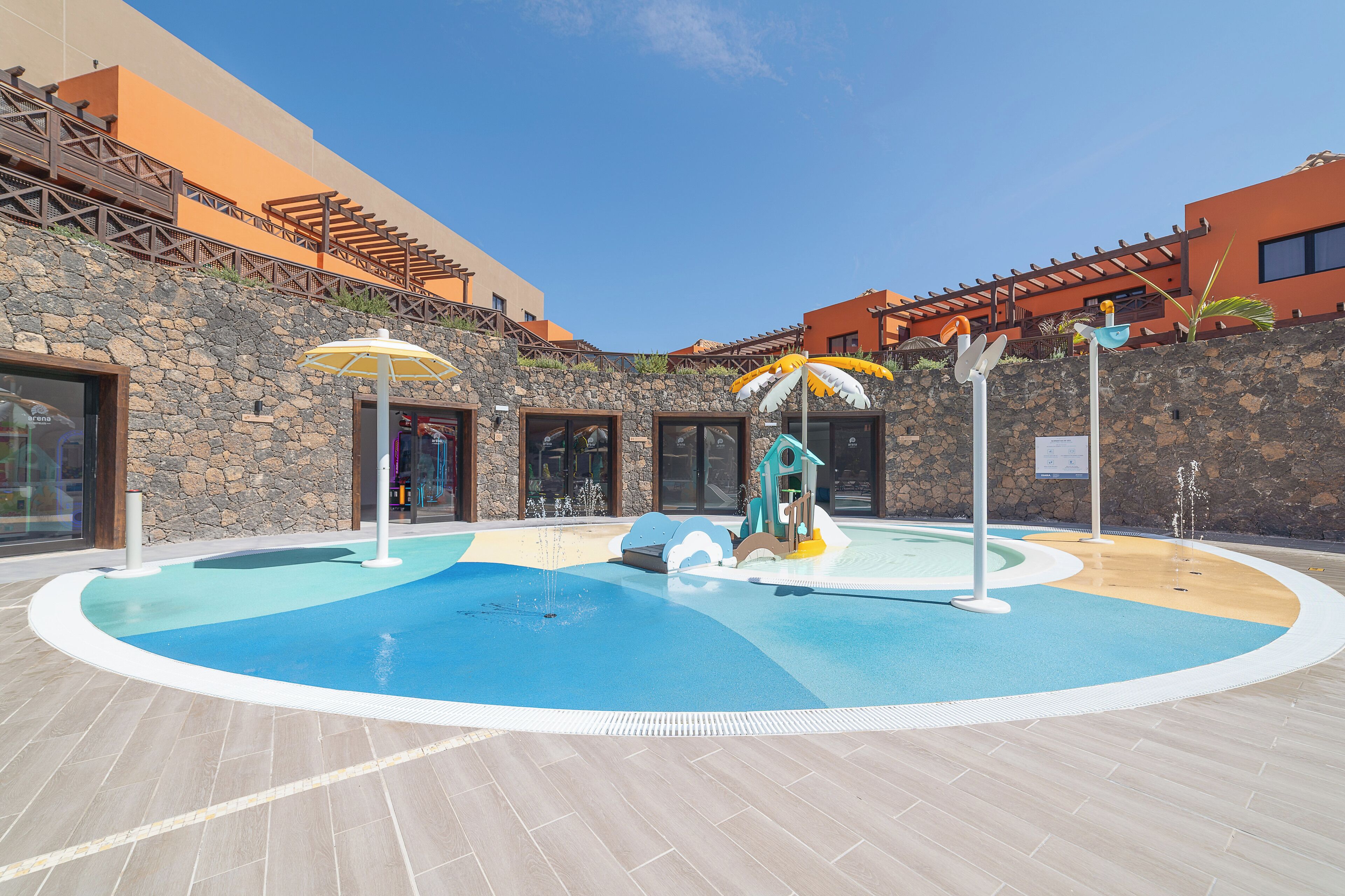 A swimming pool with a stone wall and wooden deck, surrounded by buildings with orange facades.