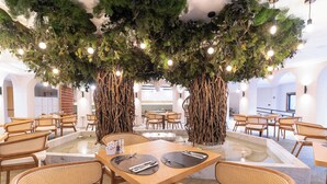 Een restaurant met ronde tafels en rieten stoelen, omgeven door een fontein en twee grote kunstbomen.