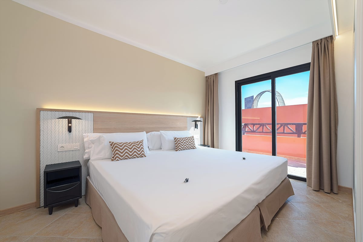 A hotel room with a large bed, a nightstand, a wall-mounted lamp, and a view of an outdoor area.