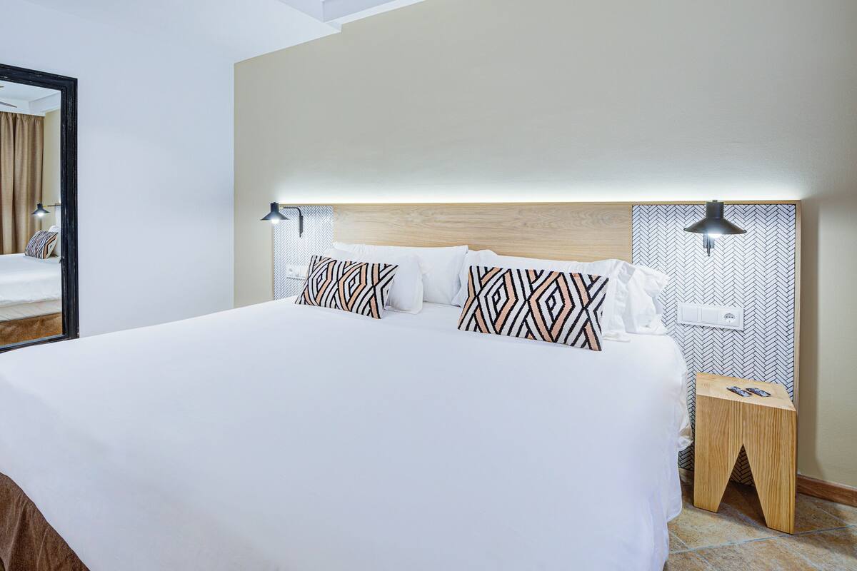 A hotel room with a large bed, a mirror, two bedside tables, and a headboard with built-in lighting.