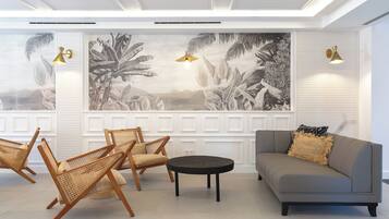 Un salon moderne avec un canapé gris, des fauteuils en osier, une table basse noire et une grande fresque tropicale sur le mur.