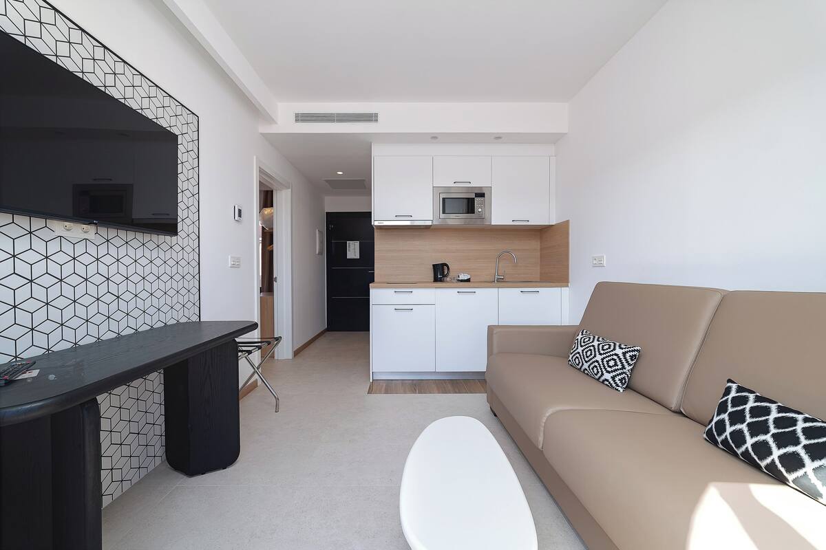 A modern kitchen with white cabinets, a microwave, and a coffee maker. There is a beige sofa with patterned pillows and a white ottoman.