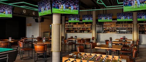 Zona de bar con varias pantallas, mesas, sillas y una mesa de fútbol.
