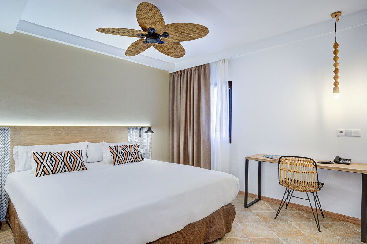 A hotel room with a large bed, a desk, a chair, a ceiling fan, and a wall-mounted light fixture.