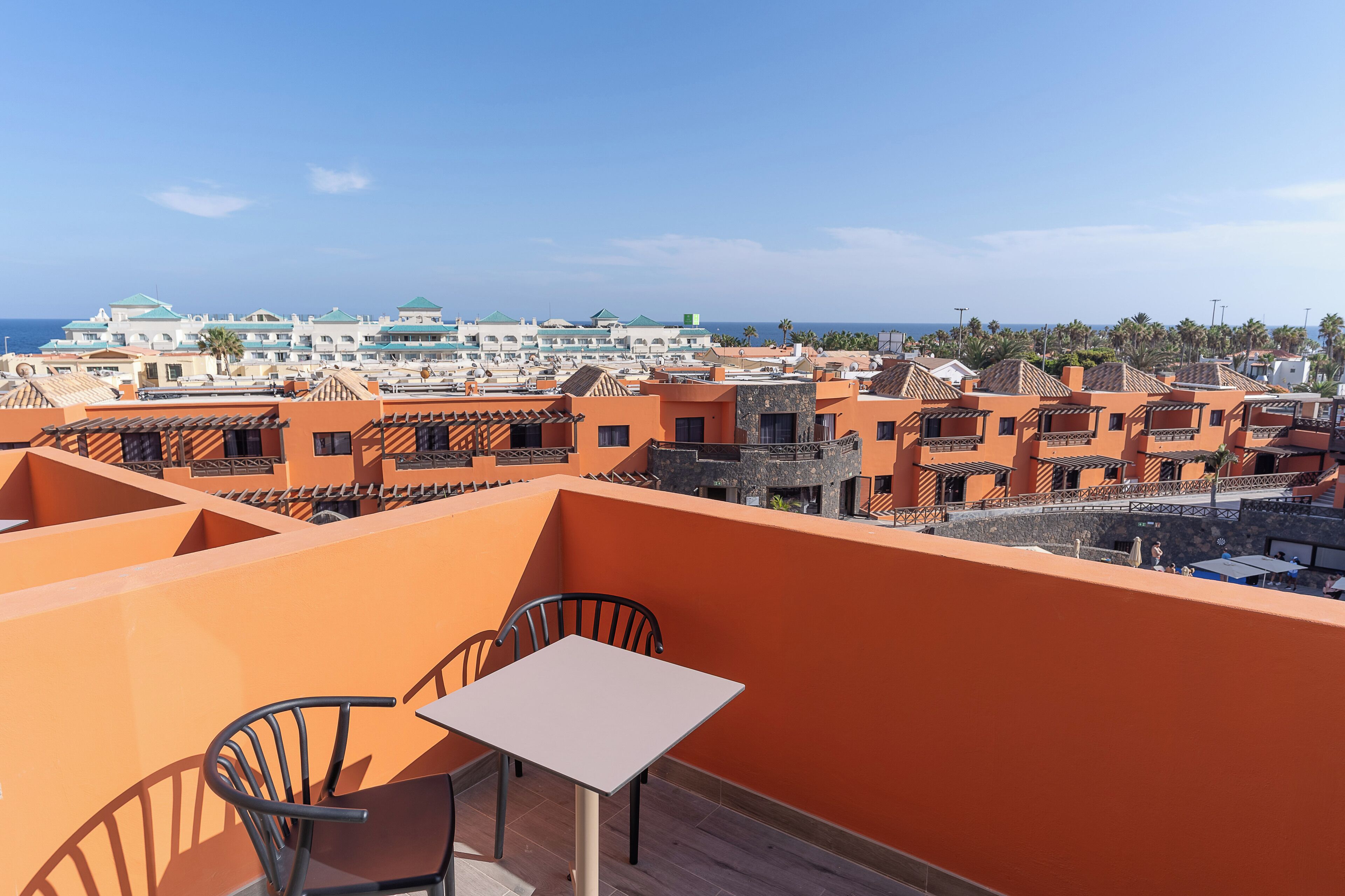 A rooftop terrace with a table and chairs, overlooking a coastal town with buildings and palm trees.