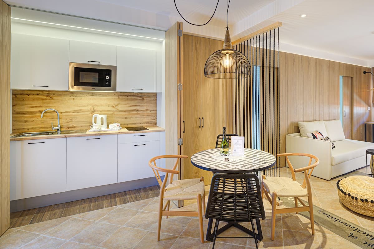 A modern kitchen with white cabinets, a built-in microwave, and a wooden backsplash. There is a dining area with a round table, two chairs, and a decorative basket. A sofa and a small side table are also visible in the living area.