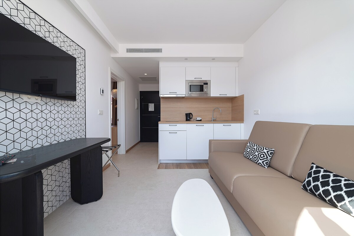 A modern kitchen with white cabinets, a microwave, and a coffee maker. There is a beige sofa with patterned pillows and a white ottoman.