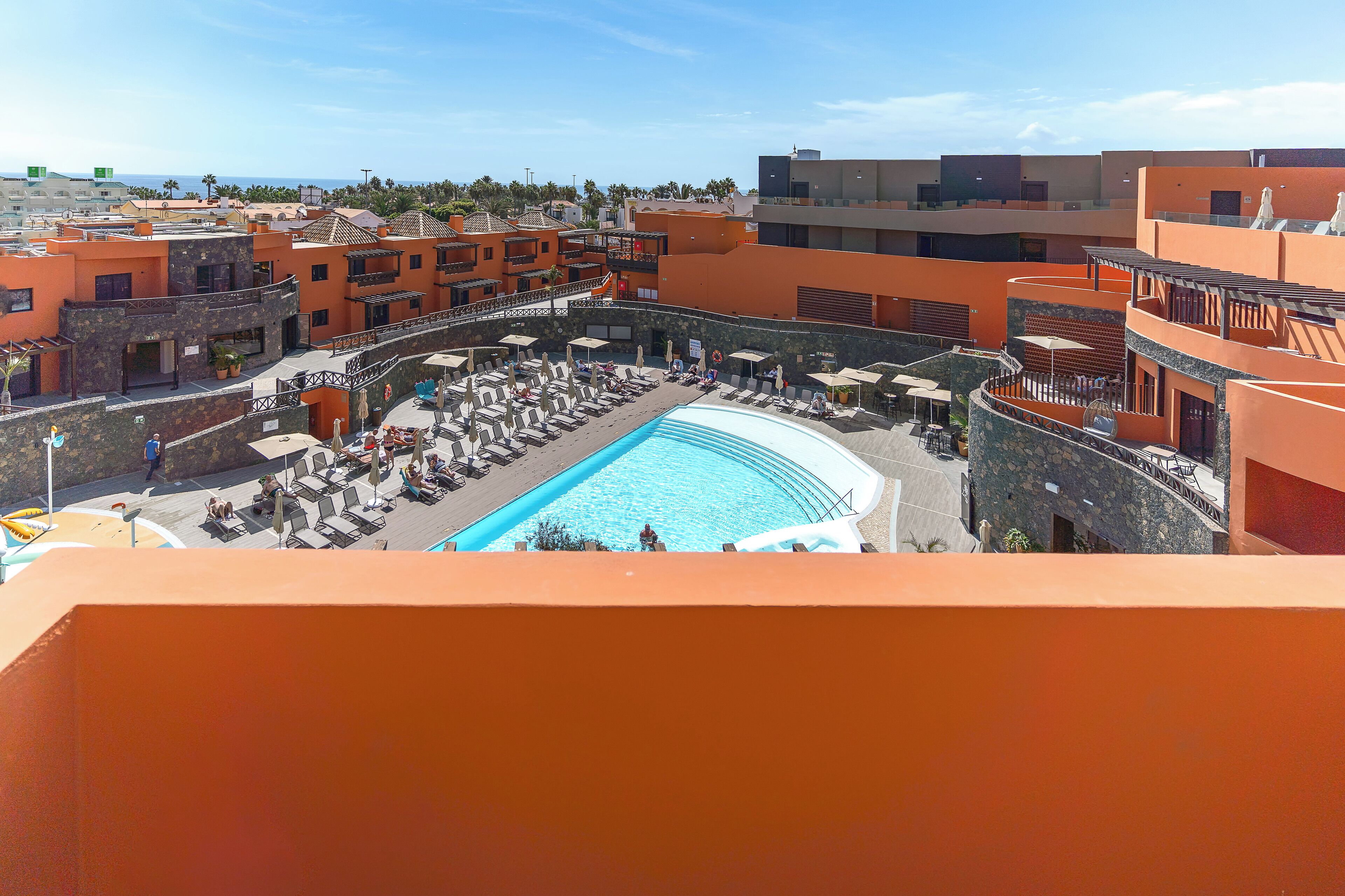 Zona de piscina en la azotea con sillones de descanso y sombrillas, rodeada de edificios anaranjados con balcones.