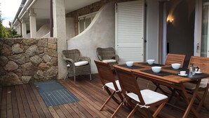 Une terrasse extérieure avec un mur en pierre, des chaises en osier, une table en bois avec des bols, et une porte dotée d’un panneau de verre.