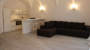 Un salon moderne avec un canapé d’angle marron foncé, un petit comptoir de bar blanc et un coin cuisine doté de meubles blancs et d’une cuisinière.
