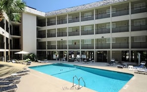 A multi-story hotel with a swimming pool, outdoor seating, and a palm tree.
