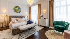 Une chambre à coucher avec une tête de lit capitonnée, un grand lit, un fauteuil vert, une commode blanche et un lustre.