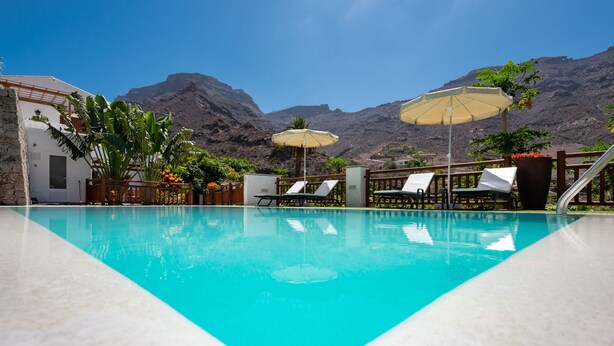 Una piscina con lettini e ombrelloni, immersa nel verde e con montagne sullo sfondo.