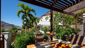 Tavola imbandita per la colazione, con frutta e un bicchiere di succo, affacciata su un paesaggio montuoso e un edificio bianco.