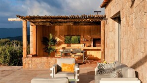 Een terras buiten met een houten pergola, een stenen muur en een zithoek met twee stoelen en een bank.