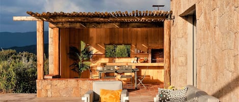An outdoor patio with a wooden pergola, a stone wall, and a seating area with two chairs and a sofa.