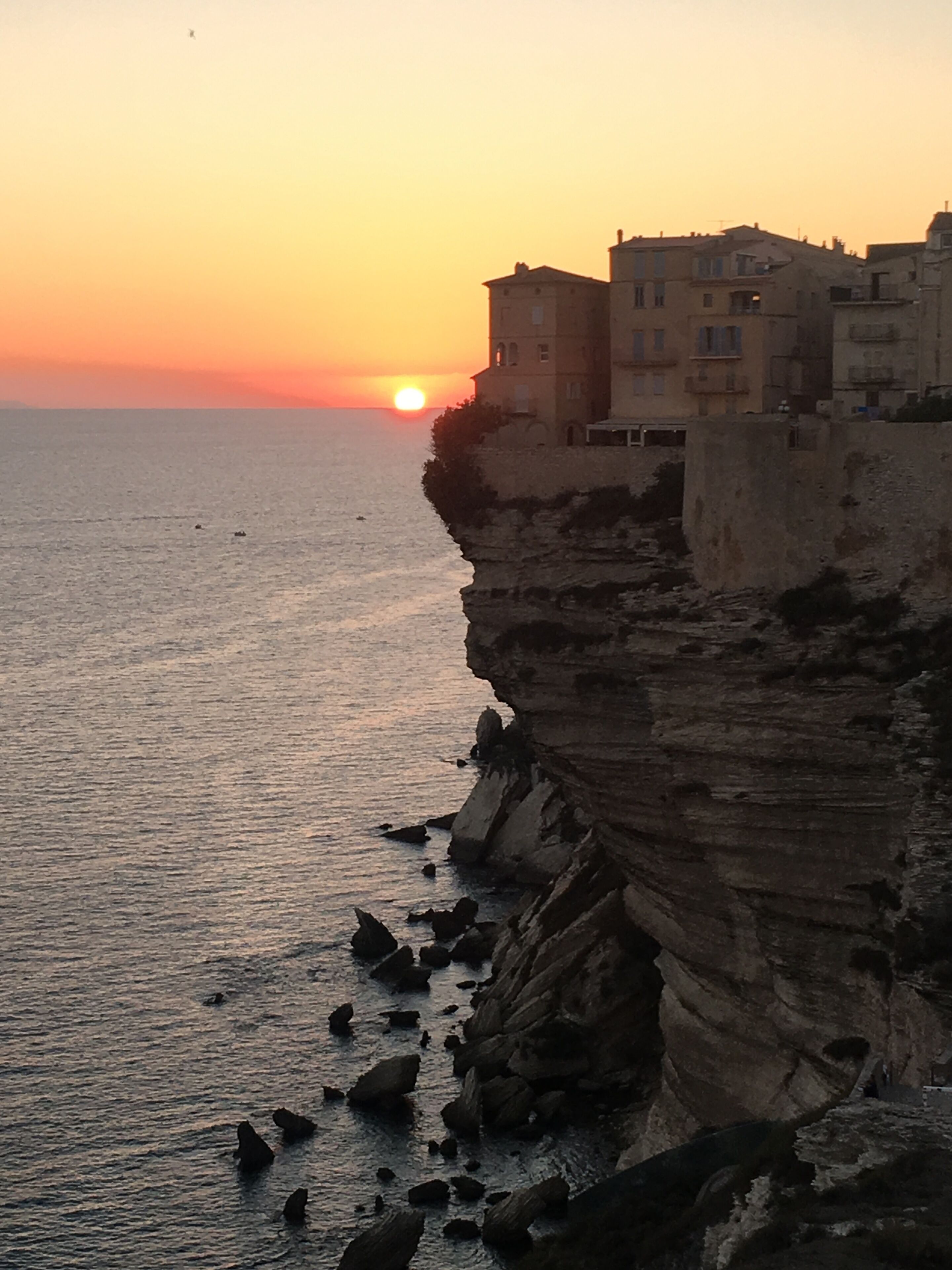 A coastal scene with a sunset, rocky cliffs, and buildings along the edge.