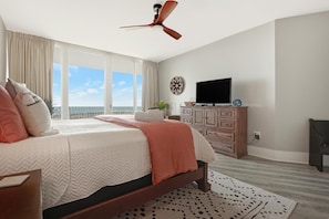 A bedroom with a bed, a ceiling fan, a TV, a dresser, and a view of the sky.
