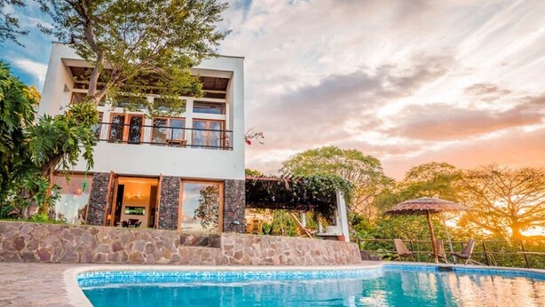 Une maison moderne à deux étages, dotée d’une piscine, de murs en pierre et d’un balcon donnant sur la piscine.