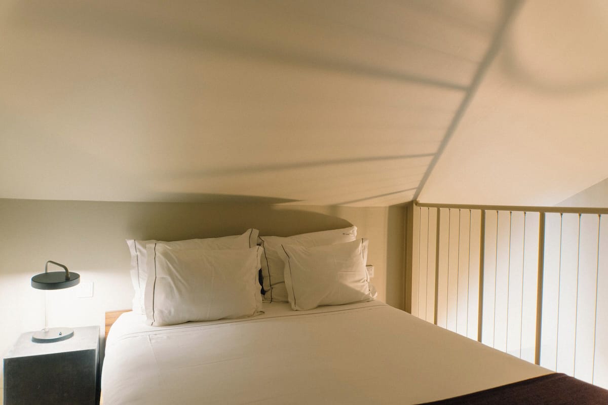 Ein ordentlich bezogenes Bett mit weißen Leinen und Kissen, ein Nachttisch mit Lampe sowie ein Fenster mit Jalousien.